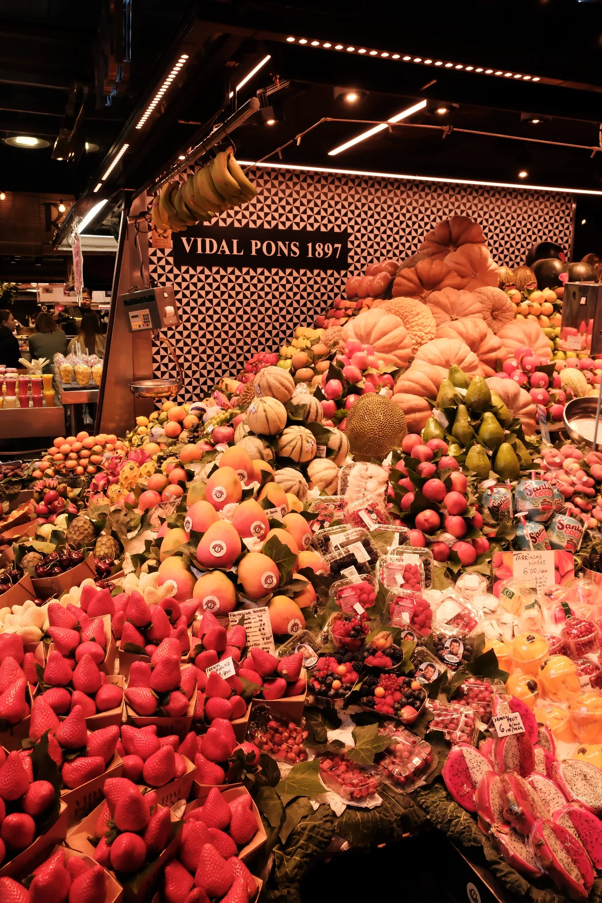 A vivid fruit stall filled with strawberries, citrus, dragon fruit, melons, and tropical produce