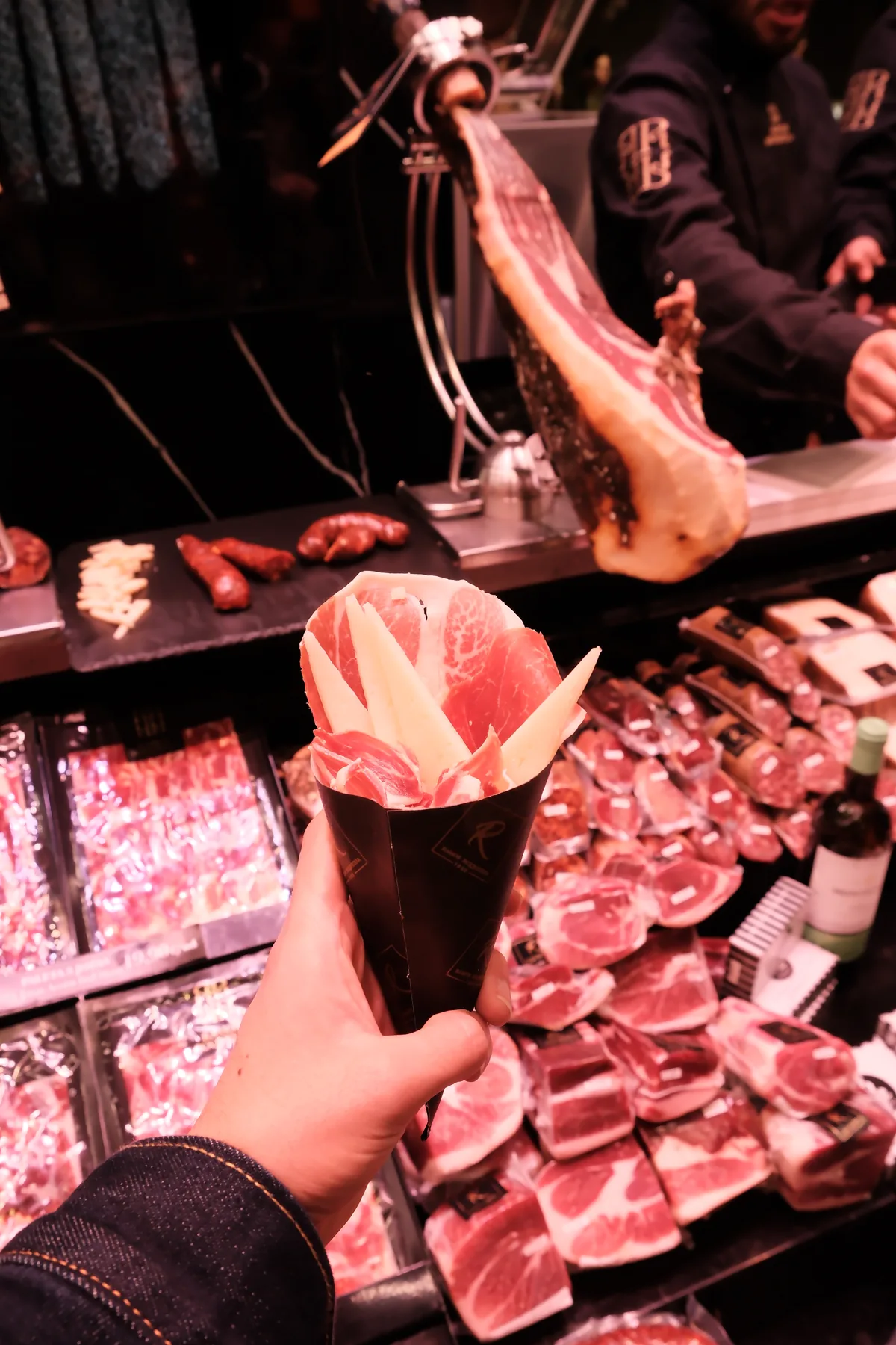 A hand holding a paper cone of sliced jamón in front of a market charcuterie counter