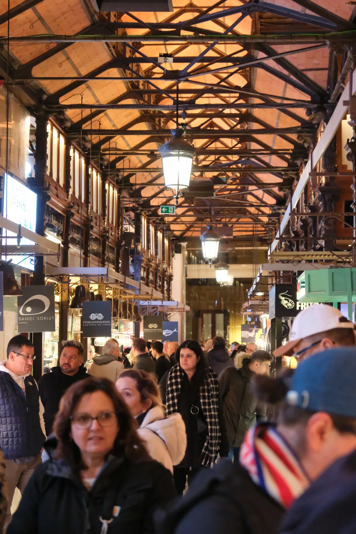 A busy aisle beneath the iron trusses and warm hanging lights of Mercado de San Miguel