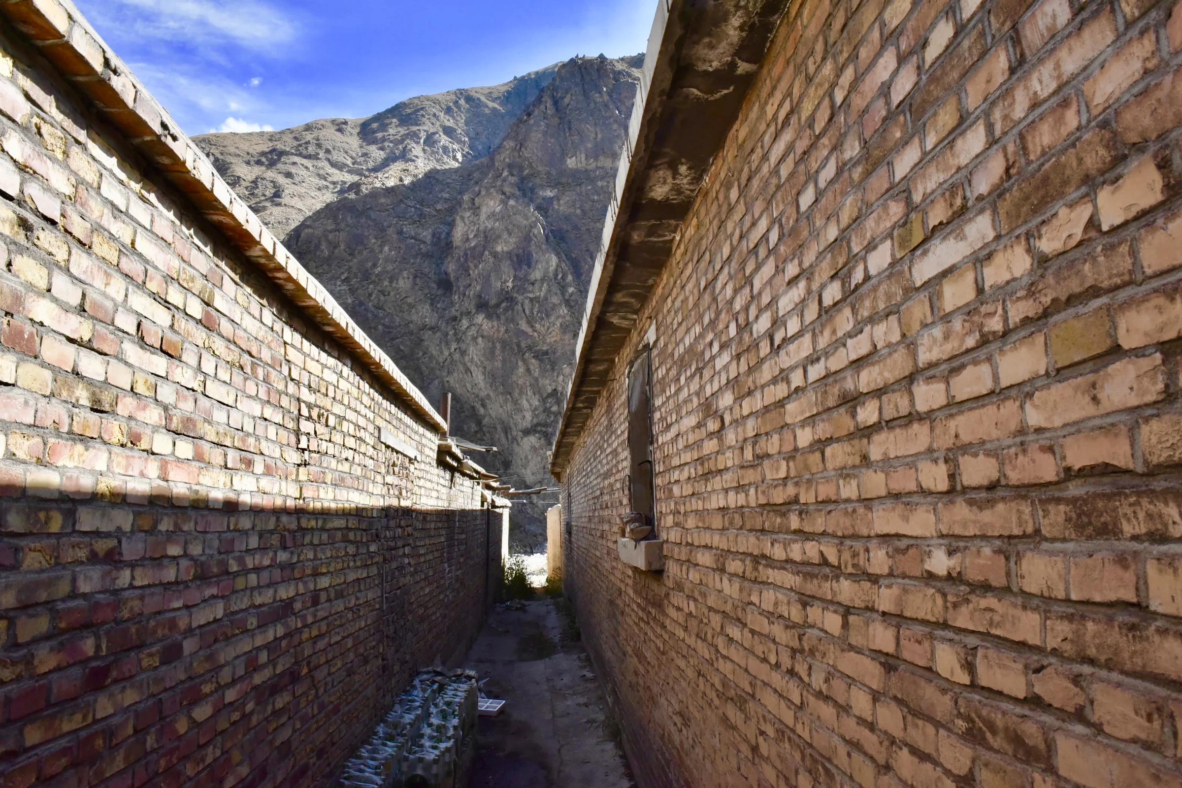 A narrow sunlit passage between long brick walls with mountains beyond
