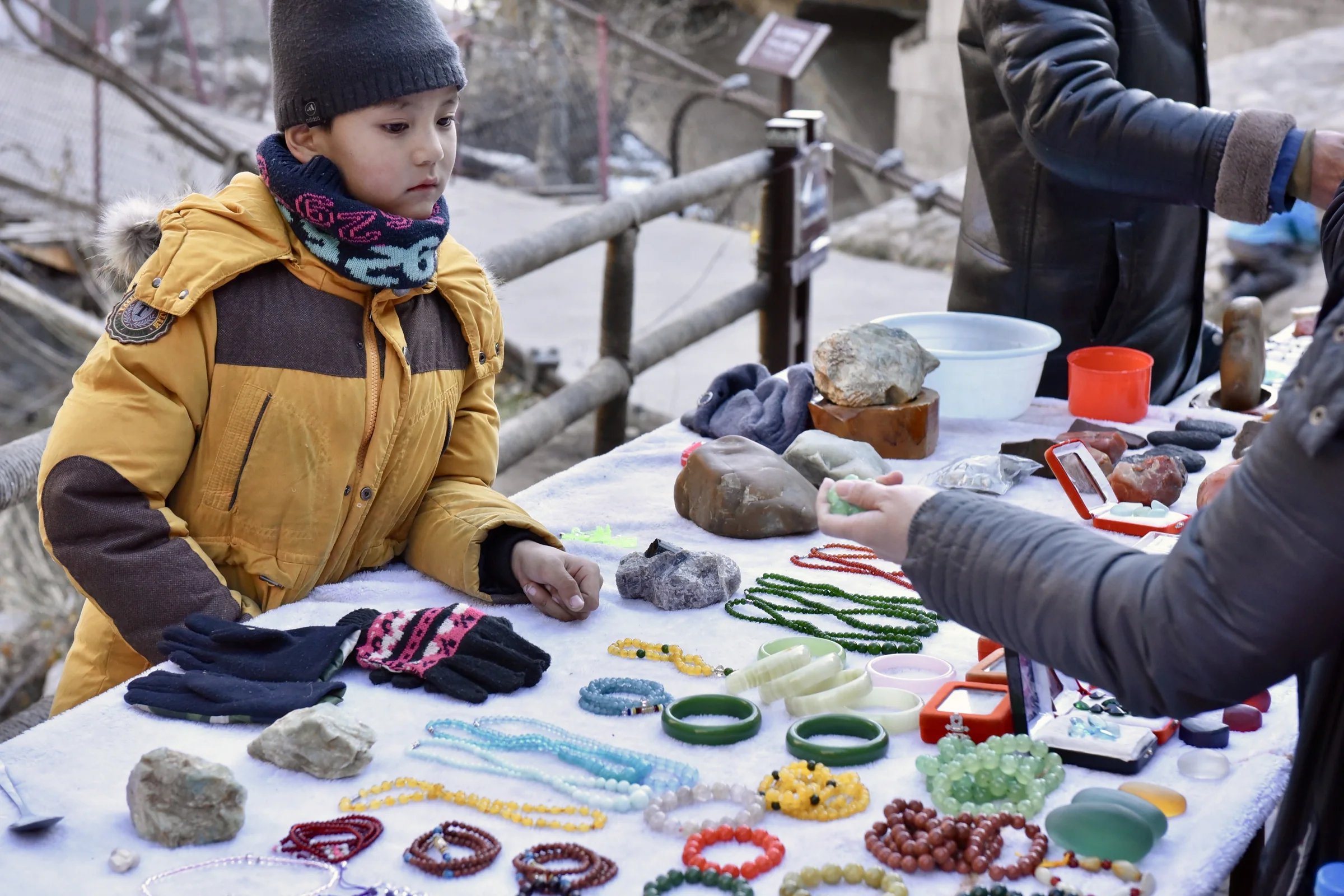 A child in a yellow coat standing beside stones and bracelets at an outdoor market